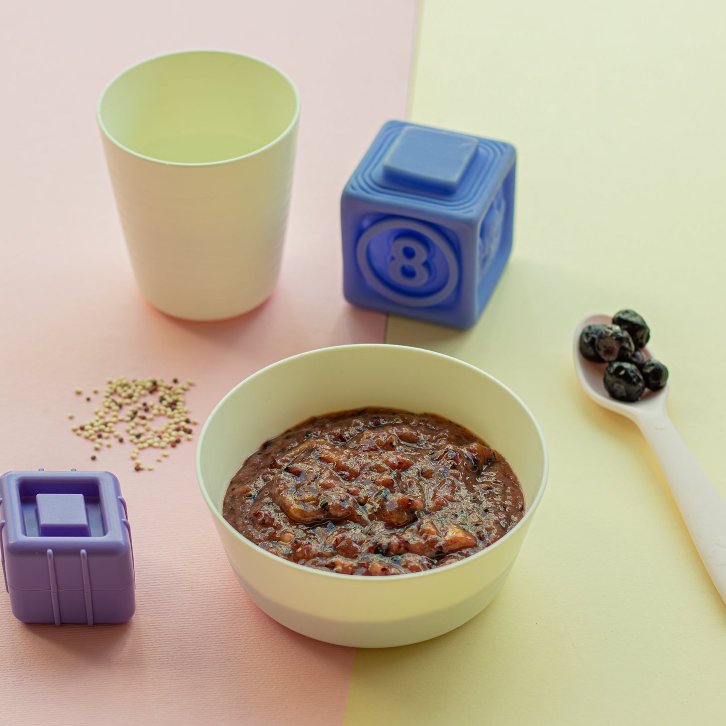 A bowl of Tropical Quinoa baby food with mixed berries and mango, alongside a spoon and containers with a pacifier design.