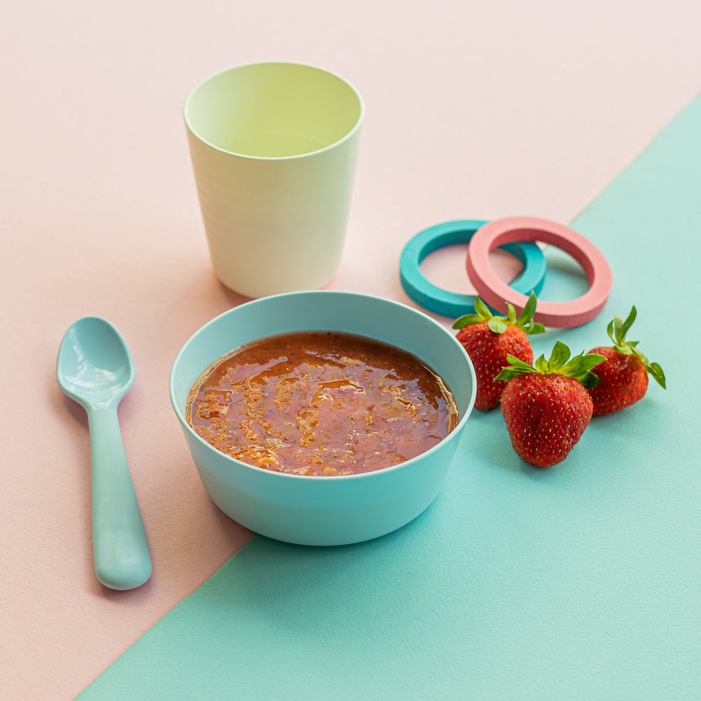 A bowl of pureed food next to a spoon and fresh strawberries, with a cup and baby feeding accessories in the background.