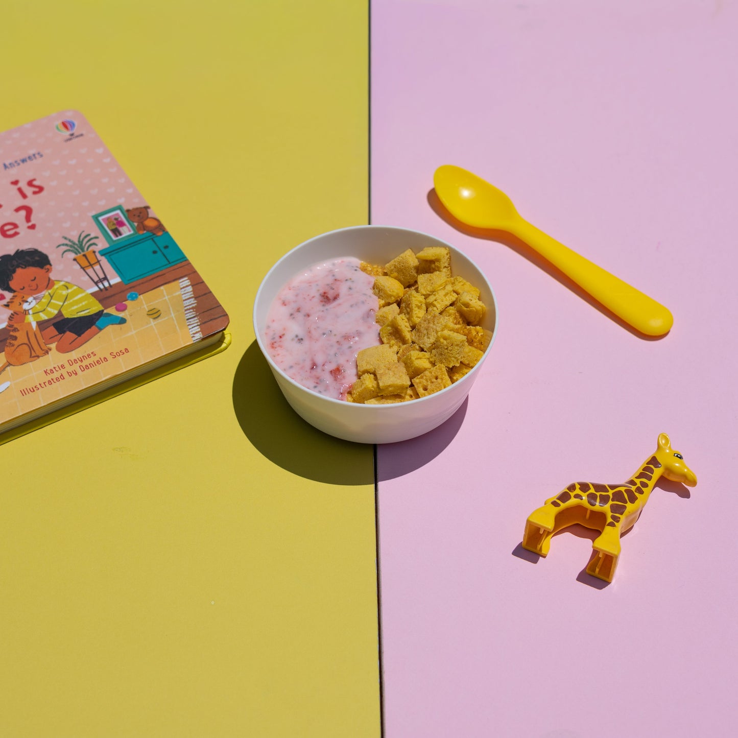 Children's book titled 'What is Love?' with a bowl of cereal, yellow spoon, and giraffe toy on a two-tone yellow and pink background.