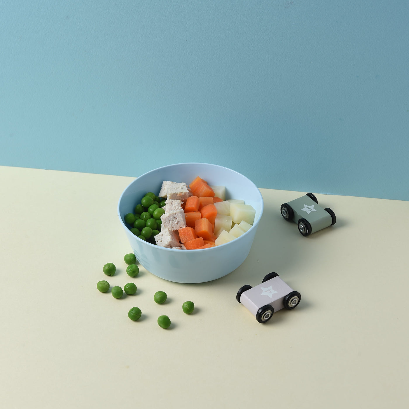 A top-down view of a light blue oval bowl filled with mixed vegetables (green peas, white cauliflower, orange carrots, and pale yellow potato) on a split cream and light blue background, with small toy cars nearby.