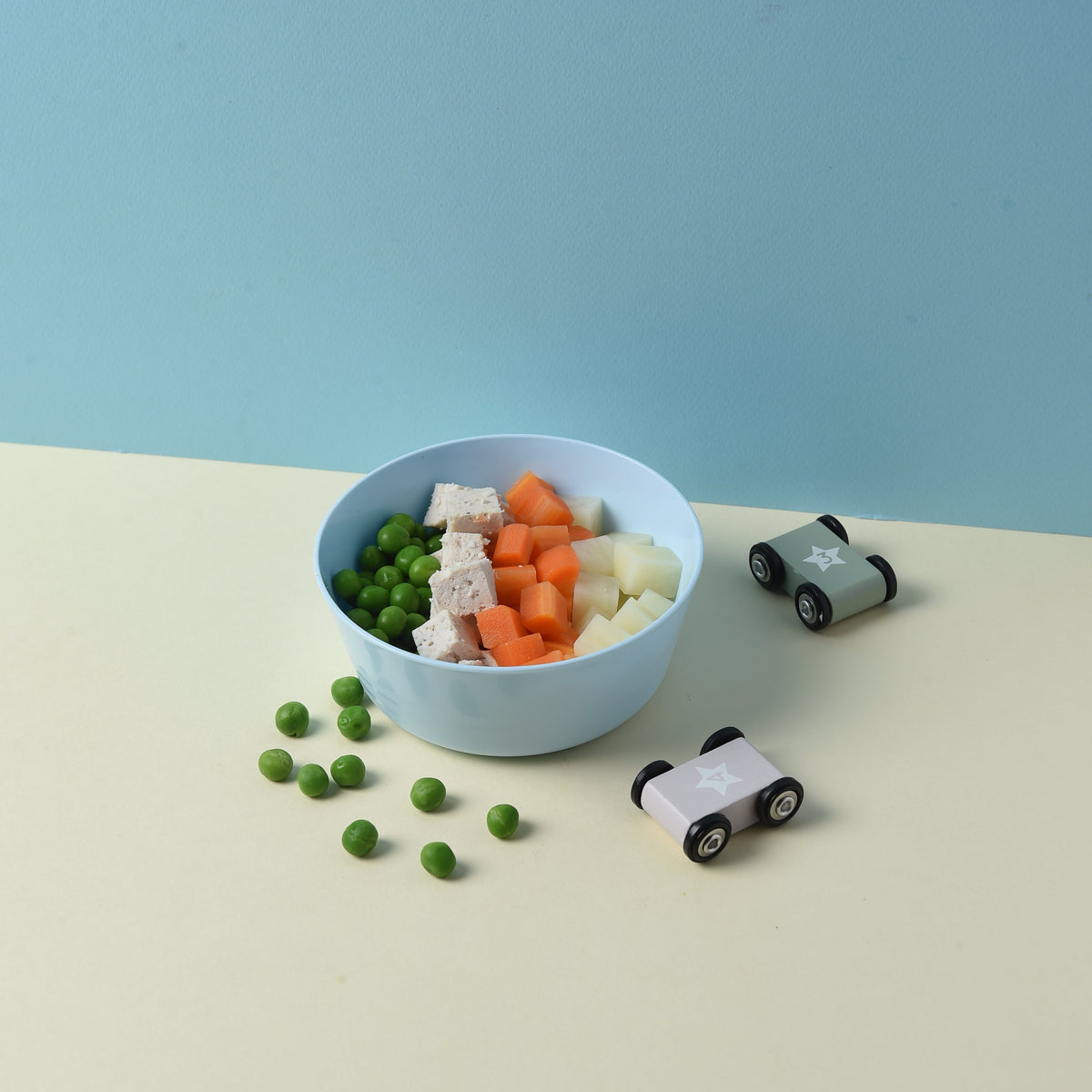 A top-down view of a light blue oval bowl filled with mixed vegetables (green peas, white cauliflower, orange carrots, and pale yellow potato) on a split cream and light blue background, with small toy cars nearby.