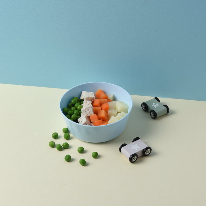 A top-down view of a light blue oval bowl filled with mixed vegetables (green peas, white cauliflower, orange carrots, and pale yellow potato) on a split cream and light blue background, with small toy cars nearby.