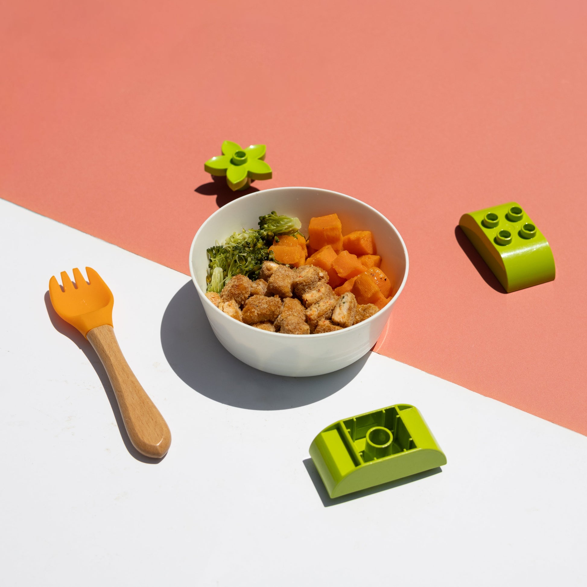 Children's bowl with food, fork, and green toy blocks on a pink and white background