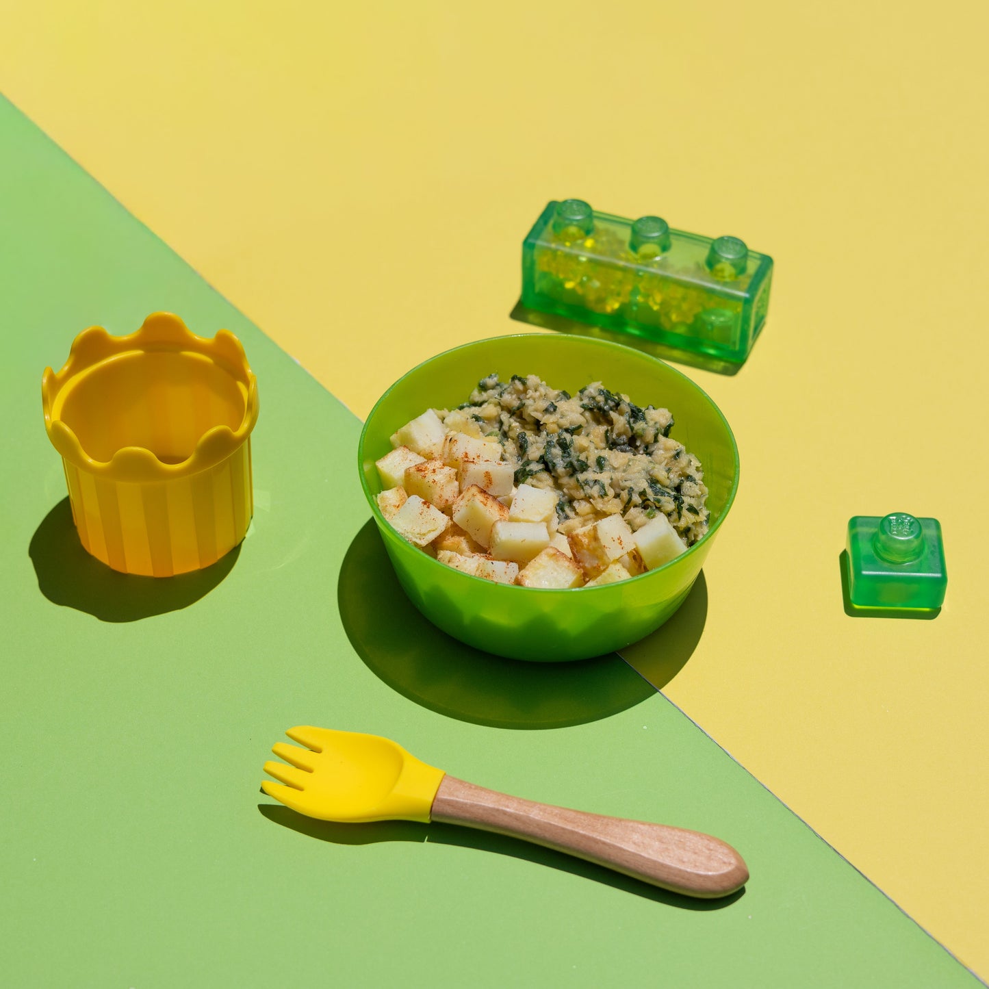 A children's meal prep set arranged on a split yellow and green background, featuring a green bowl filled with food, a yellow utensil with a wooden handle, a yellow cup, and two translucent green toy-like pieces.
