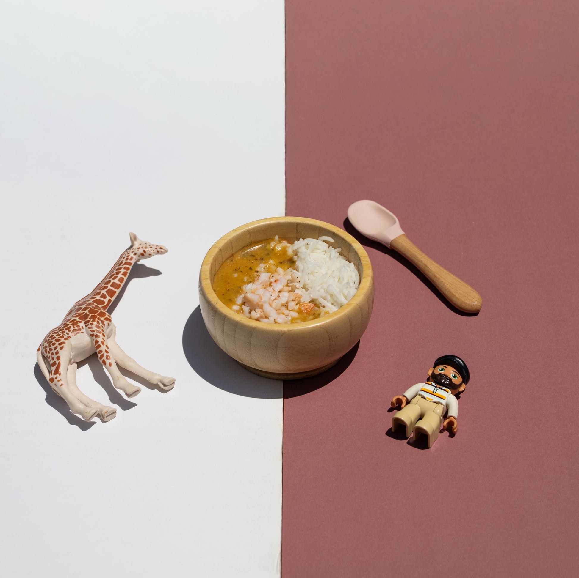 A styled flat-lay featuring a small wooden bowl of rice and yellow-orange soup, a wooden spoon, a giraffe figurine, and a small child figurine, arranged on a split white and dusty rose background.