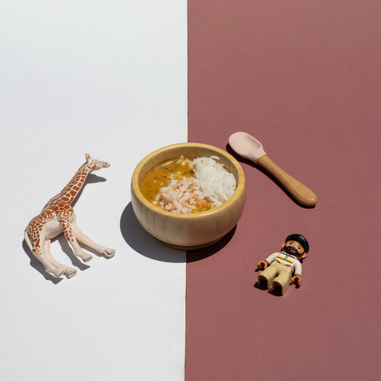 A styled flat-lay featuring a small wooden bowl of rice and yellow-orange soup, a wooden spoon, a giraffe figurine, and a small child figurine, arranged on a split white and dusty rose background.